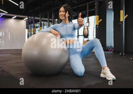 Sportivo soddisfatto in un abbigliamento sportivo blu si siede sul pavimento della palestra con una palla fitness e mostra il suo pollice in su Foto Stock