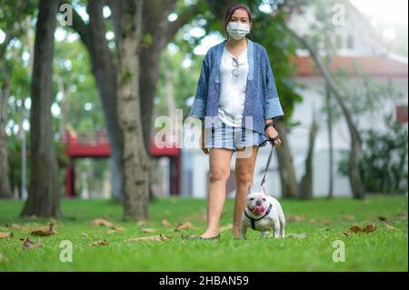 Donna con bulldog francese sul prato, donna in maschera medica in piedi e un bulldog francese bianco con un guinzaglio, New Normal. Foto Stock