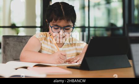 Bambina asiatica con occhiali che fanno i suoi compiti in un notebook con un laptop davanti. Foto Stock