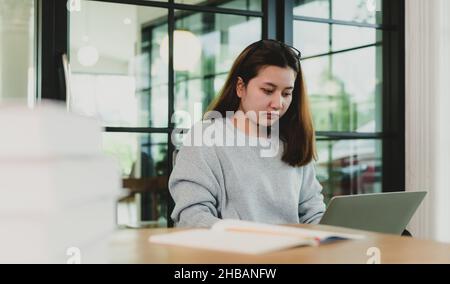 Una donna adolescente piega la testa per azionare un computer portatile posto sulla gamba, lavora a casa guardando serio. Foto Stock