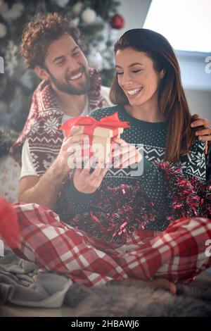 Un ragazzo sta dando un regalo di natale alla sua ragazza amico mentre sono seduti sul pavimento in una piacevole atmosfera a casa insieme. Natale, casa, Foto Stock