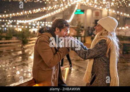 Un ragazzo giovane sta riscaldando le mani della sua ragazza mentre camminano sotto le luci di natale in una bella notte di neve in città. Natale, Capodanno, Foto Stock