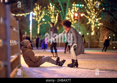 Una ragazza sta aiutando il suo ragazzo a levarsi in piedi mentre impara a pattinare alla pista di pattinaggio su ghiaccio durante le vacanze di natale in una bella notte in città. CH Foto Stock