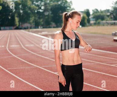 La giovane ragazza allegra in abbigliamento sportivo guarda lo smart watch all'aperto in uno stadio rivestito di rosso Foto Stock