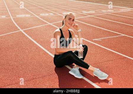 Donna Happy Fit in abbigliamento sportivo che ascolta la musica con le cuffie e tiene la bottiglia d'acqua mentre si siede su una pista da stadio con rivestimento rosso all'aperto. Esegui Foto Stock