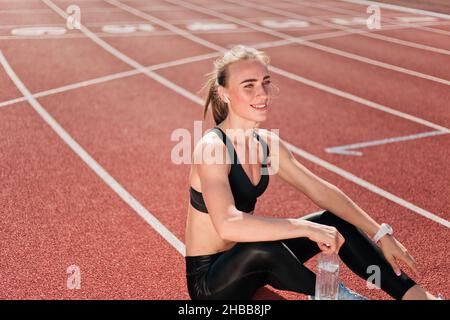 Donna Happy Fit in abbigliamento sportivo che ascolta la musica con le cuffie e tiene la bottiglia d'acqua mentre si siede su una pista da stadio con rivestimento rosso all'aperto. Esegui Foto Stock
