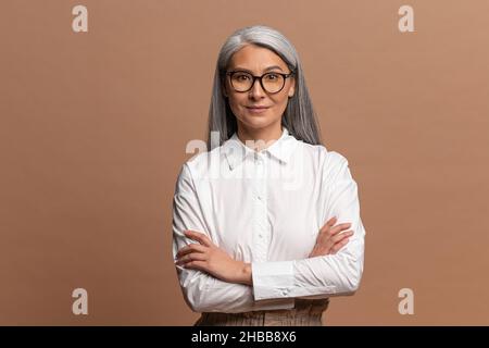 Ritratto di bella positiva vecchia donna in piedi con le braccia incrociate, aspetto gentile e sorriso affascinante. Studio interno girato isolato su sfondo beige Foto Stock
