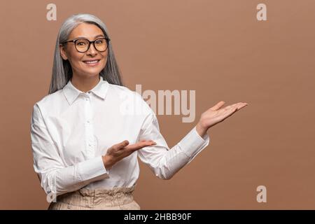 Donna anziana positiva puntando palme via con il sorriso toothy sulla faccia, spazio vuoto per la vostra pubblicità, indossando vestito ufficiale di stile. Studio interno girato isolato su sfondo beige Foto Stock
