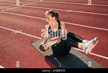 La giovane donna in forma perfezionerà il suo corpo mentre fa l'esercitazione con la sfera della medicina sul tappeto sul tracciato dello stadio con rivestimento rosso alla giornata luminosa di sole. Sana vita Foto Stock