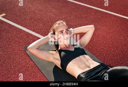 Giovane donna in forma perfezionerà il suo corpo facendo esercizio di torsione per i muscoli addominali sul tappeto al percorso dello stadio con rivestimento rosso in giorno di sole luminoso. Sano l Foto Stock