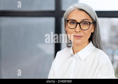 Vista ritratto di donna anziana, sicura e sorridente, che indossa occhiali guardando la fotocamera con un sorriso di piacere. Concetto di lavoratore dipendente seduto in ufficio Foto Stock