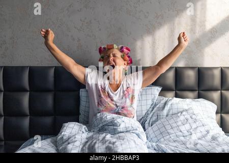 Donna di mezza età che sbadigia presto al mattino coperto con coperta bianca sul letto in camera da letto di casa. Sogni dolci, Buongiorno, nuovo giorno, fine settimana, Foto Stock
