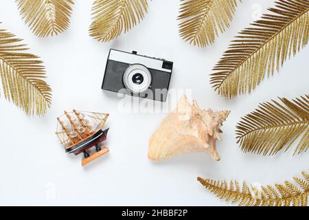 Viaggio minimalista ancora vita. Seashells, fotocamera retrò tra decorativi foglie di palma dorate su sfondo bianco. Vista dall'alto Foto Stock