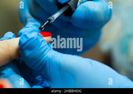 Un manicurista in guanti blu, coprendo le unghie con una pasta abrasiva a lunga durata in gel rosso. Foto Stock