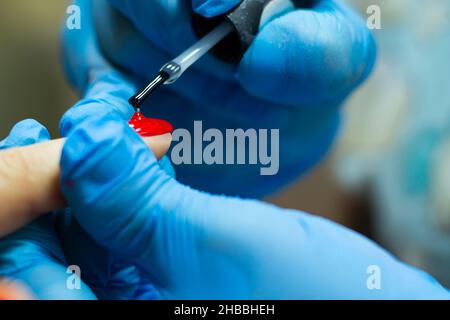 Un manicurista in guanti blu, coprendo le unghie con una pasta abrasiva a lunga durata in gel rosso. Foto Stock