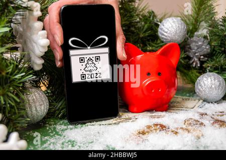 Una mano che tiene uno smartphone con un simbolo regalo e un codice QR a forma di abete, una banca di maialino rosso su monete d'oro coperte di neve e Christma Foto Stock