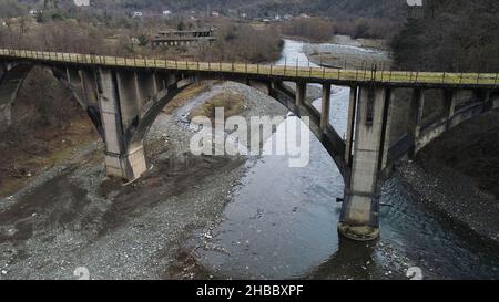 Volare sotto il ponte di cemento distrutto e rotto sullo sfondo della foresta autunnale e un fiume con riva di pietra. Antenna di montagna fredda st Foto Stock