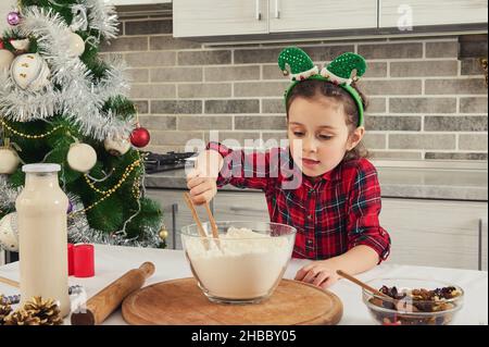 Adorabile ragazza con l'elfo di cerchio sulla testa mescola accuratamente ingredienti secchi con farina in ciotola di vetro in piedi sul tavolo accanto all'albero di Natale. Li Foto Stock