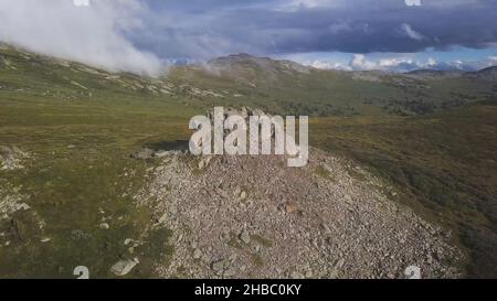 Vista aerea su prato verde con solitario sfondo roccioso e cielo nuvoloso. Paesaggio di montagna incredibile con grande roccia al centro dell'erba. Antenna Foto Stock