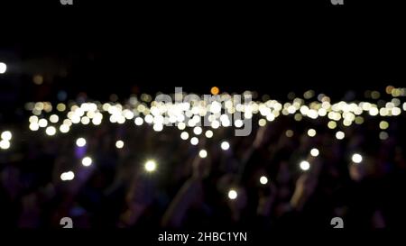 Spettatori sfocati in sala con luci che ondolano al buio. Pubblico che tiene i telefoni con le luci scintillanti durante il concerto, atmosfera romantica. Foto Stock