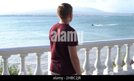 Uomo biondo che cammina lungo la passeggiata in una città del sud vicino al mare. Giovane ragazzo pensivo che cammina lungo il mare e le montagne blu ondulato, concetto di summe Foto Stock