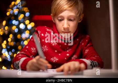 Un ragazzo in un maglione rosso di Natale sullo sfondo di un albero di Natale tiene un marcatore in mano e si disegna su carta Foto Stock