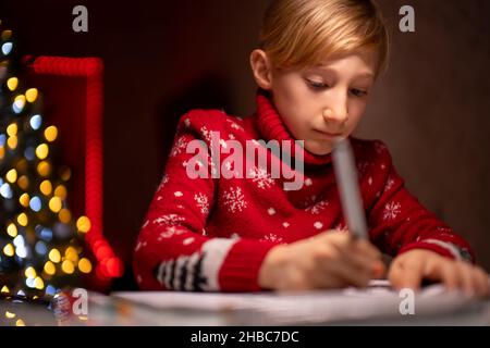 Un ragazzo in un maglione rosso di Natale sullo sfondo di un albero di Natale tiene un marcatore in mano e si disegna su carta Foto Stock