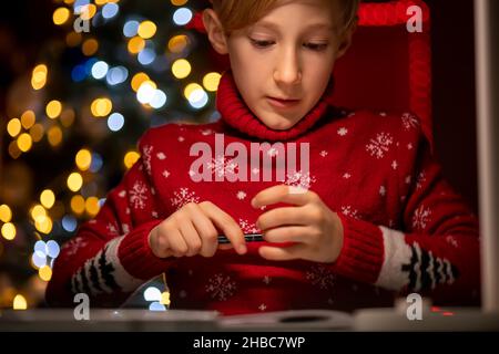 Un ragazzo in un maglione rosso di Natale sullo sfondo di un albero di Natale tiene un marcatore per il disegno Foto Stock