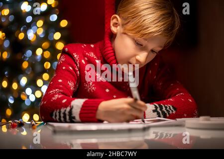 Un ragazzo in un maglione rosso di Natale sullo sfondo di un albero di Natale tiene un marcatore per il disegno Foto Stock