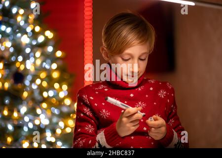 Un ragazzo in un maglione rosso di Natale sullo sfondo di un albero di Natale tiene un marcatore per il disegno Foto Stock