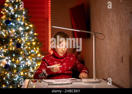 Un ragazzo in un maglione rosso di Natale sullo sfondo di un albero di Natale illuminato da una lampada tiene in mano un disegnatore Foto Stock