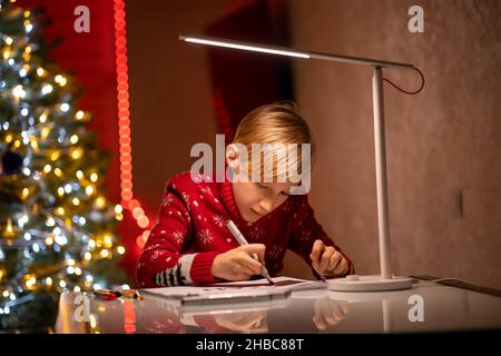 Un ragazzo in un maglione rosso di Natale sullo sfondo di un albero di Natale illuminato da una lampada tiene in mano un disegnatore Foto Stock