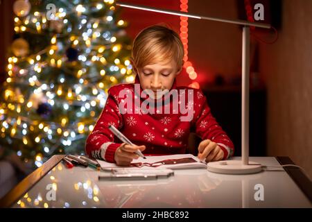 Un ragazzo in un maglione rosso di Natale sullo sfondo di un albero di Natale illuminato da una lampada tiene in mano un disegnatore Foto Stock