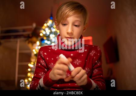 Un ragazzo in un maglione rosso di Natale sullo sfondo di un albero di Natale tiene un marcatore davanti a lui e vuole aprirlo Foto Stock