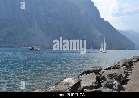 Paesaggio del Lago di Como a Lecco in estate con rocce in primo piano, barche a motore e barche a vela in movimento. Foto Stock