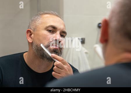 Un bell'uomo di mezza età che gli spazzolava la barba la mattina in bagno guardando nello specchio Foto Stock