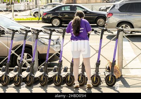 Vista posteriore di una dipendente femminile che organizza lo scooter Urent parcheggiato in una fila di scooter pick-up sul marciapiede stradale nella strada di San Pietroburgo, Russia Foto Stock