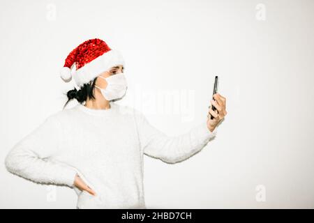 Giovane donna con facemask su tiene il cellulare alto isolato su sfondo bianco. Concetto di facetime e cummunicazione durante le vacanze su pandemia. Foto Stock