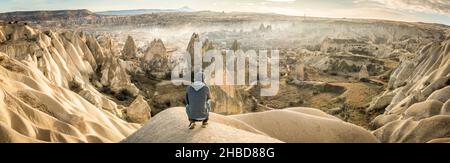 Uomo basso in ginocchio che guarda sopra la valle drammatica con Goreme città sullo sfondo. Esplorazione solista in Turchia. Viaggio cinematografico di destina Foto Stock