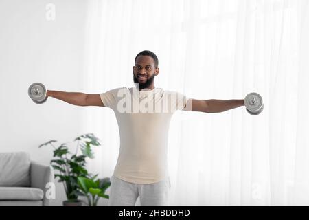 Felice bell'atleta millennial nero bearded uomo in bianco abbigliamento sportivo facendo esercizio a mano con dumbbell Foto Stock
