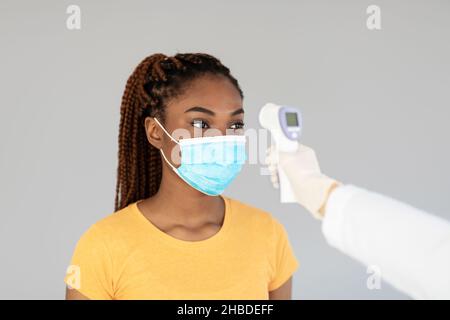 Giovane donna nera in maschera facciale che controlla la temperatura corporea con termometro elettronico su sfondo grigio Foto Stock