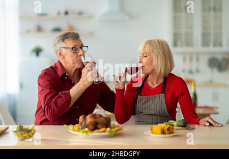 Affettuosa coppia anziana bere vino rosso mentre si prepara la cena natalizia, facendo arrostito tacchino in cucina Foto Stock