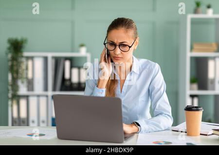 Donna d'affari arrabbiata che parla sullo smartphone e guarda il computer, avendo una conversazione seria con il cliente Foto Stock