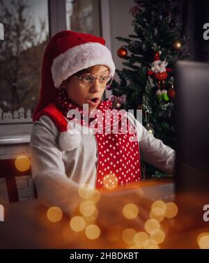 Divertente ragazzo domandandosi seduto di fronte allo schermo del portatile con cappuccio rosso di Babbo Natale che ha incontri di Natale con parenti o amici. Luci di festa. Foto Stock