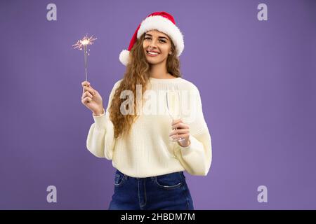 Sorridente Santa girl indossa un maglione bianco che tiene un bicchiere di champagne e scintillante su sfondo viola Foto Stock