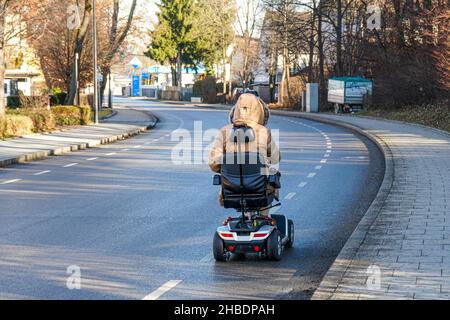 Una donna disabile sta guidando la sua sedia a rotelle elettrica su una pista ciclabile accanto alla strada. Foto Stock