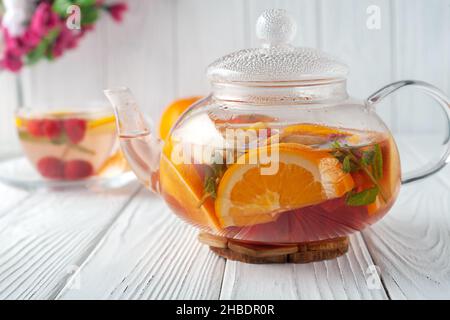 Tè alla frutta con bacche, arance e menta in una teiera di vetro su sfondo bianco di legno Foto Stock