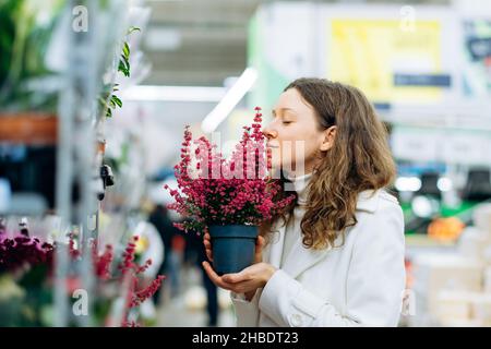 Donna con capelli ricci brunette in caldo maglione e cappotto bianco odore fiori viola in fiore in pentola con occhi chiusi in piedi in grande magazzino Foto Stock