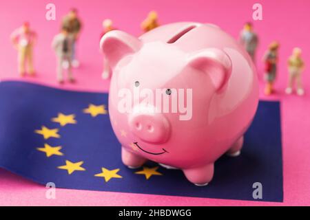 Piggy bank, bandiera dell'UE e giocattoli di plastica su sfondo colorato, un concetto sul tema del reddito della popolazione dell'Unione europea Foto Stock