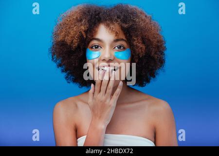 La giovane donna afroamericana con capelli corti marroni ricci e macchie blu sulle guance sorride ampiamente e tocca la bocca contro il closeup parete blu Foto Stock